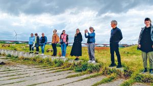 people standing in a line on concrete blocks in a grassy landscape with a distant bridge and wind turbine